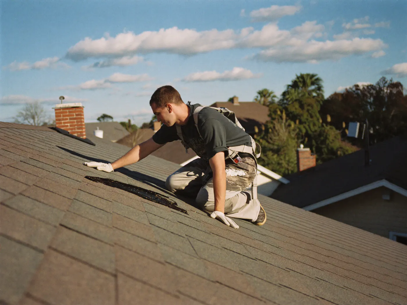A roofer kneels on an asphalt-shingle roof, inspecting damaged shingles.